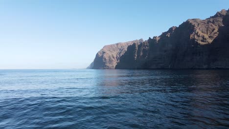 Drone-shot-ascending-from-black-sand-shore-towards-rugged-landscape-of-sea-cliffs-and-blue-ocean-water-in-Los-Gigantes,-Tenerife,-Canary-Islands