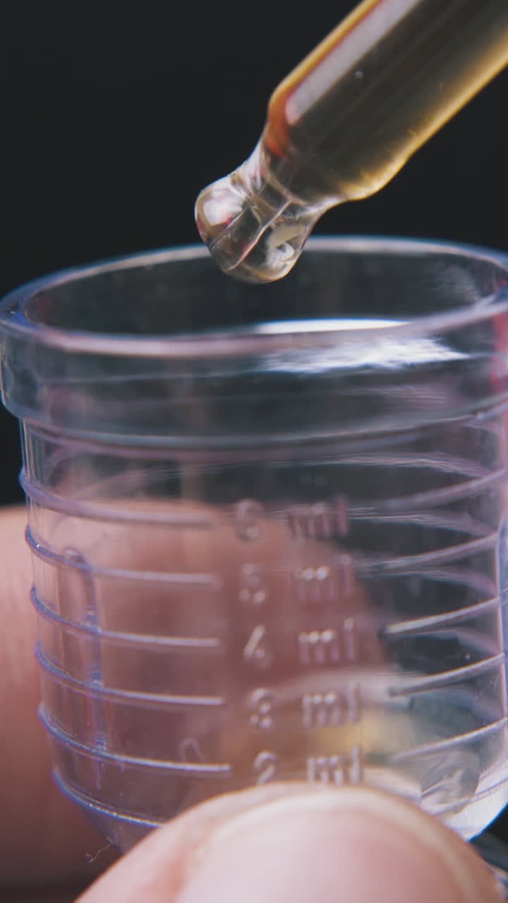scientist drops light brown liquid from pipette into plastic measuring cup on black background extreme close view