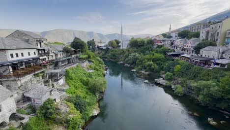 wide-angle view of mostar riverside with beautiful stone buildings, mosques, greenery and mountains in the background on a wonderful sunny morning; taken from mostar bridge - bosnia and herzegovina