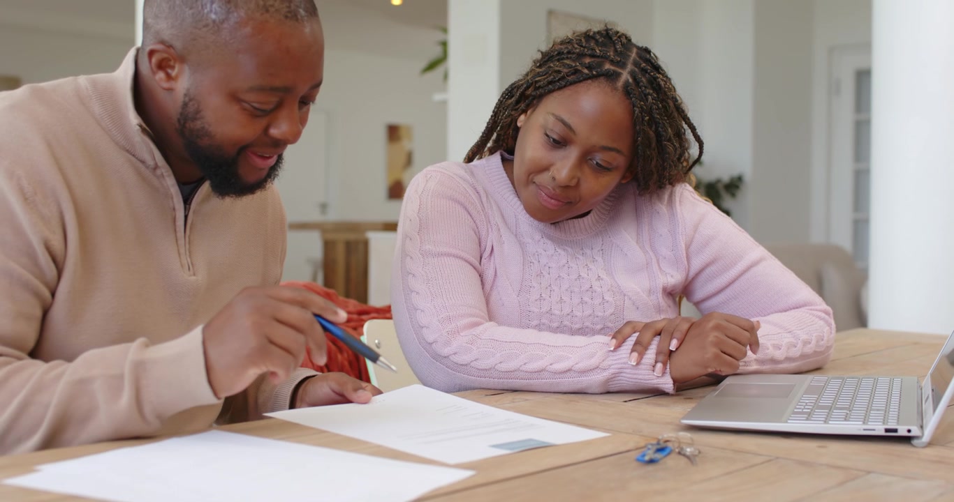 African couple sitting at wooden table man picking sheet, flipping, pointing pen, reviewing forms