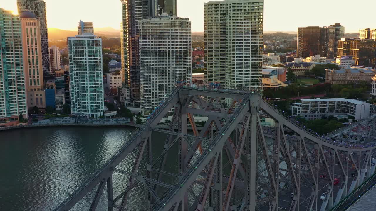Aerial flyover Brisbane River captures the traffic on Story Bridge, people climbing the cantilever bridge, and stunning views of riverside cityscape at sunset, with golden light shining over the city.