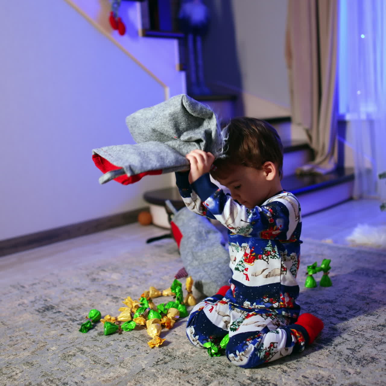 Dark-haired baby boy in winter pajama sits near Christmas tree. Lovely boy gets some candies from the stocking.