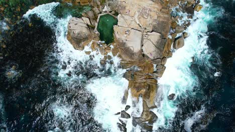 Top-down-view-of-people-at-Saunders-Rocks-Beach-as-waves-crash-onto-the-boulders,-aerial