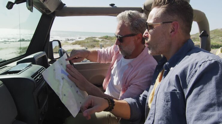 Felice coppia di uomini gay caucasici seduta in auto che legge una mappa e indica, in una giornata di sole al mare.