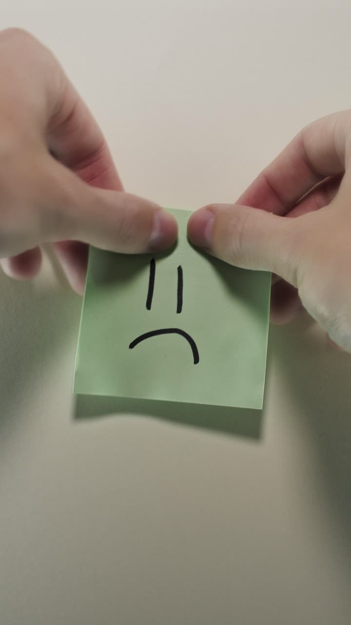 Hands posting Sad Face sticky note on light wall, evoking disappointment