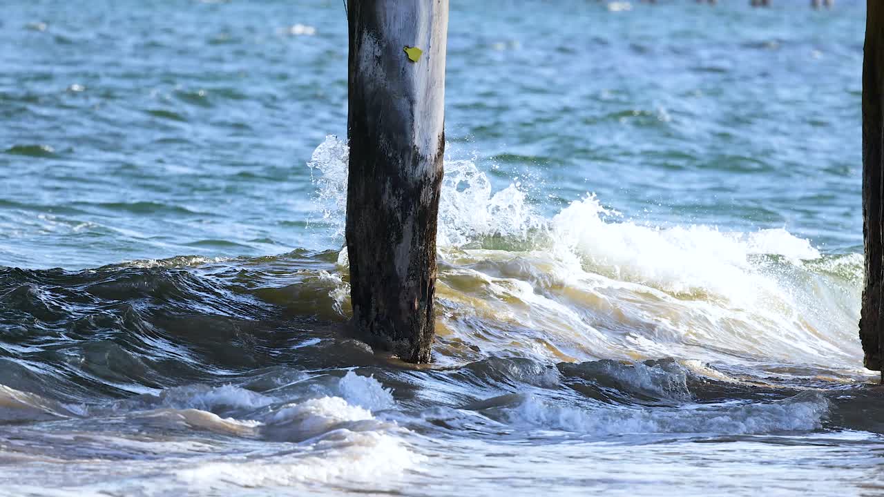 Ocean waves rhythmically crash against a wooden pier under bright daylight, capturing the dynamic interaction of water and structure
