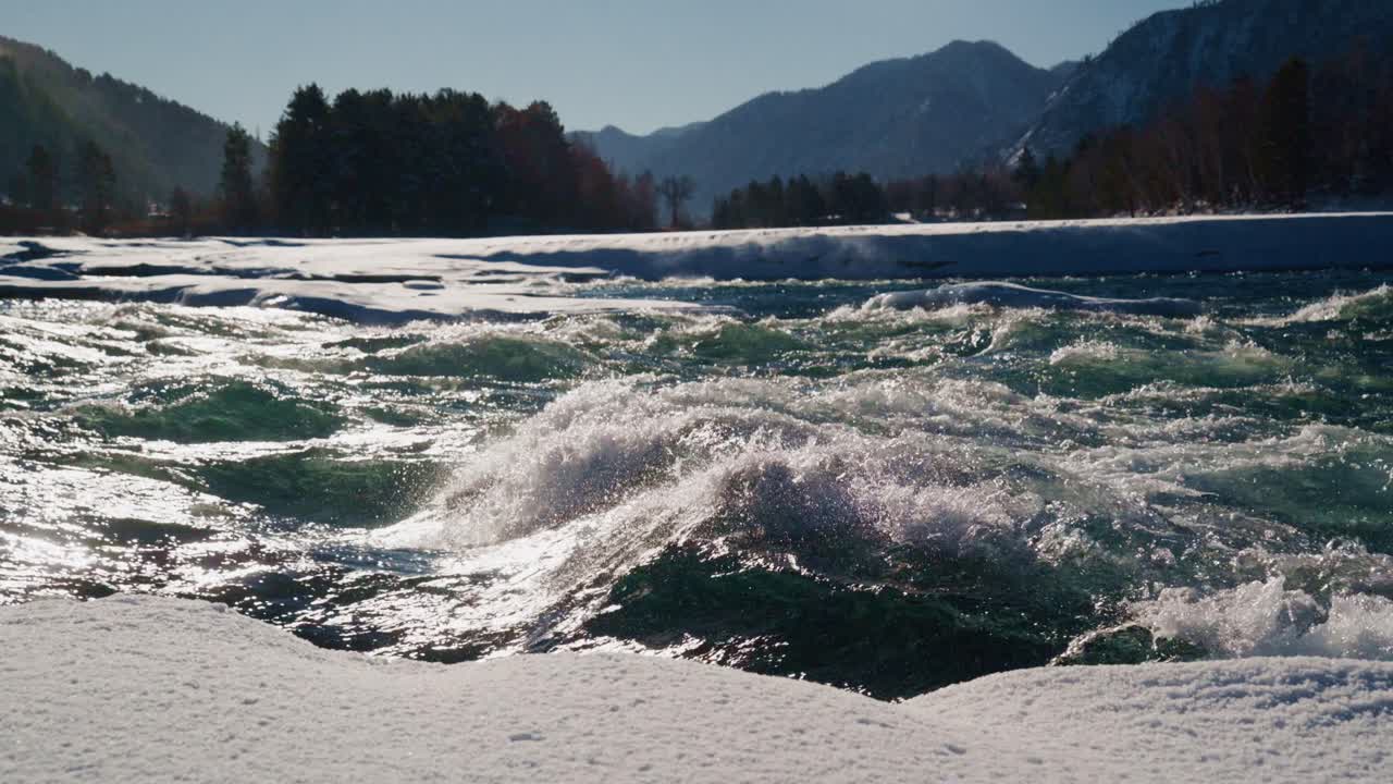 Discover the beauty of rapid waters swirling in a snowy terrain surrounded by majestic mountains. The winter sun enhances the tranquil yet dynamic nature of the scene