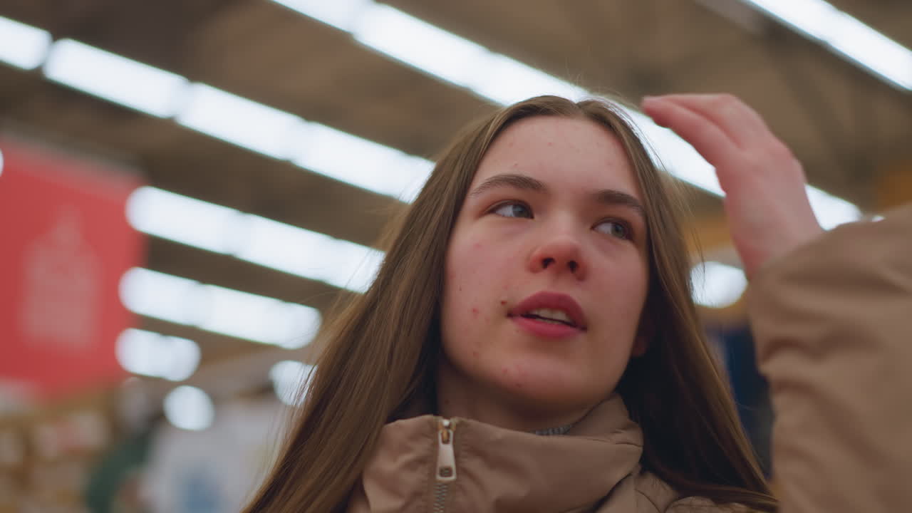 갈색 재을 입은 어린 소녀가 쇼핑몰에서 그녀의 머리카락을 부드럽게 만지는 클로즈업. 배경은 흐릿하며 소녀의 사려 깊은 표정과 캐주얼한 몸짓을 강조합니다.
