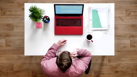 young man in glasses typing on laptop and picking up phone, topshot, sitting behind desk with coffee and documents