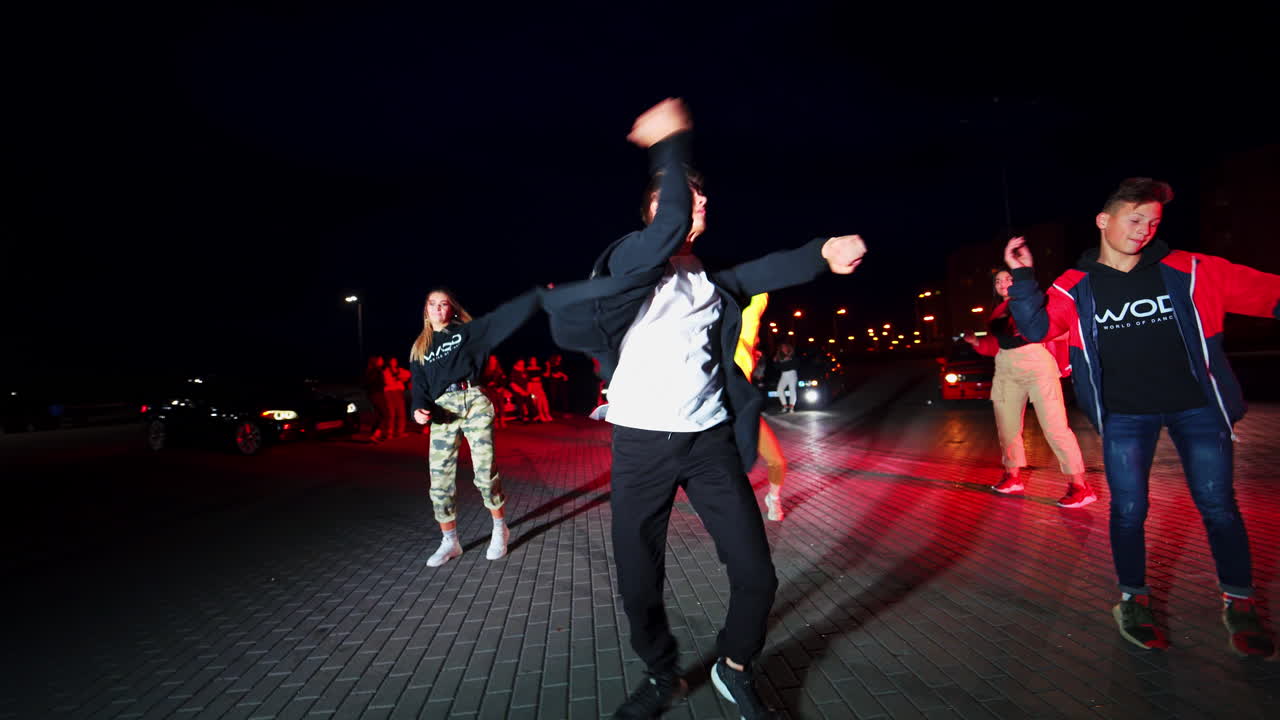 Teenage boys and girls dance in the bright flashing colorful lights outdoors at night. Crowd scene near cars at backdrop.