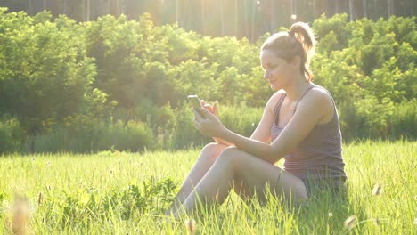 happy young woman sitting on green lawn and uses smartphone on scenic field at sunset background