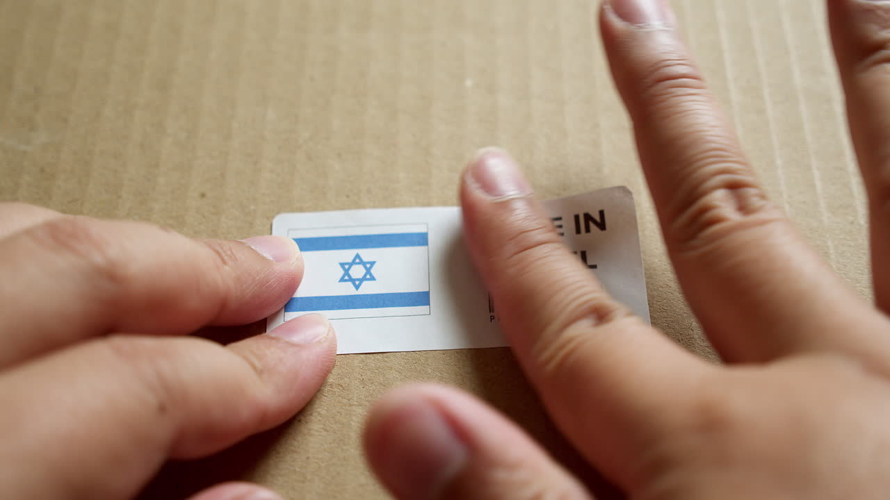 Mani che applicano un'etichetta con bandiera "MADE IN ISRAEL" su una scatola di spedizione con codice a barre di qualità premium del prodotto