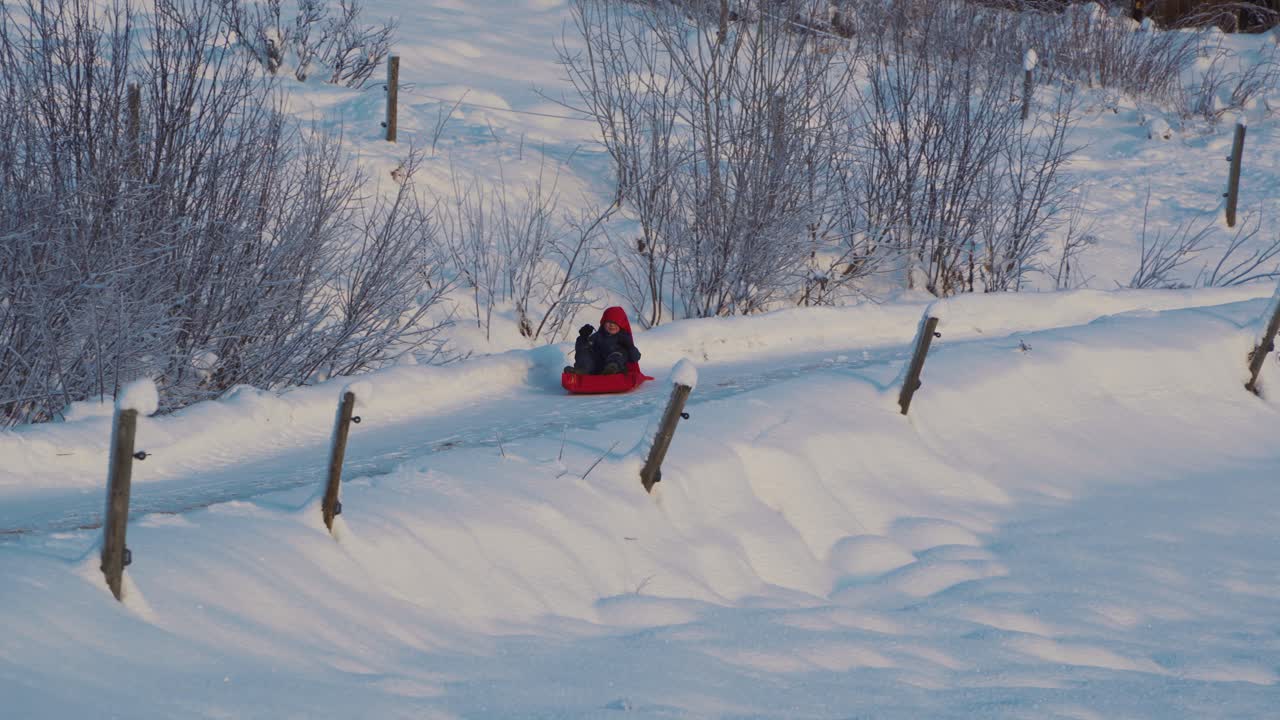 Sporingsopptak av ung gutt i kjelke som kjelker ned en snødekt vei om vinteren på en solrik dag