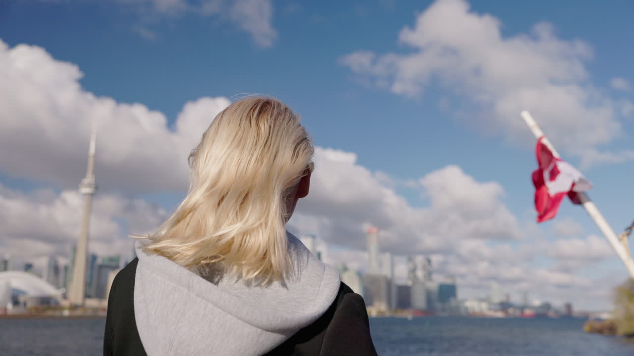 Young woman tourist looking at Toronto skyline from boat on Lake Ontario, back view with blonde hair in wind and Canadian flag