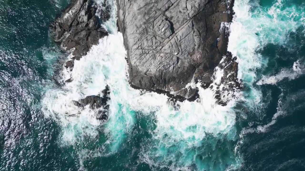 Vista a volo d'uccello di una grande formazione rocciosa piatta circondata da onde spumeggianti turchesi e spruzzi di mare bianchi contro l'oceano blu profondo.