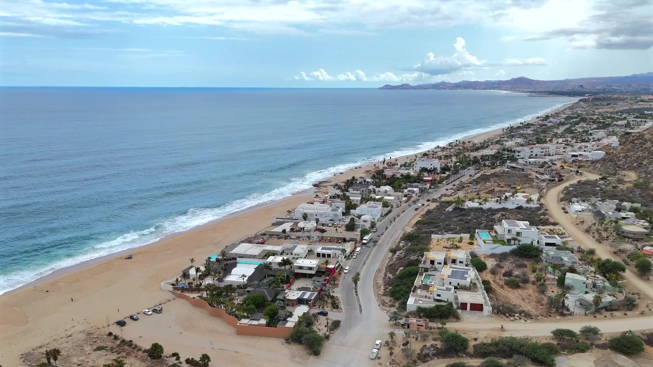 Drone avança das colinas de La Laguna em direção à praia, com a comunidade na parte inferior do quadro, a costa e o oceano no meio, e Palmilla Point e o céu acima.