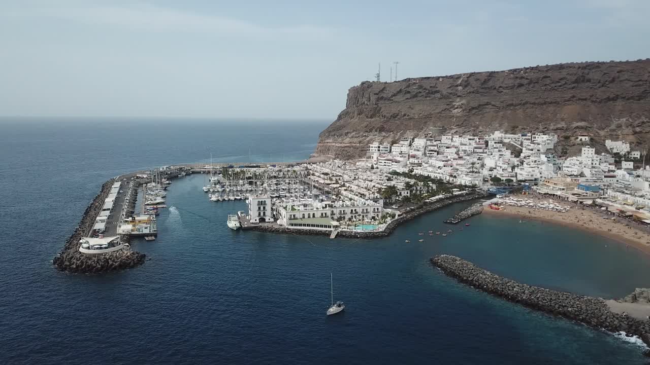 A drone flies over the turquoise waters off the coast of Puerto de Mogán, showcasing the bustling marina and port area filled with fishing boats and yachts.