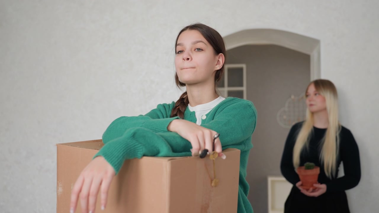 Young woman leaning on box, keys in hand, roommate with potted plant in background quiet apartment doorway, cardboard stacked, hesitant expression, paperwork and lease handover tension, lingering