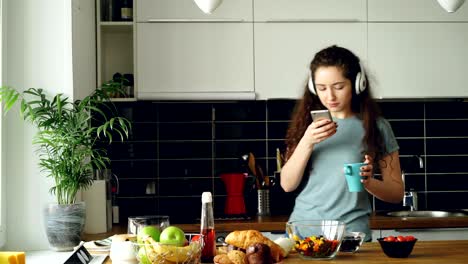 atractiva chica feliz bailando y cantando en la cocina mientras usa el teléfono inteligente y escuchando música en casa por la mañana