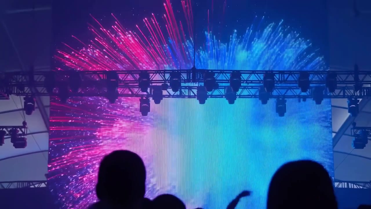 Bagfra lavvinkelbillede af silhuetpublikum, der vender mod kæmpe LED-scene med intens magenta og cyan radial glød, overhead truss-lys, levende stribeteksturer, dramatisk kontrast, fordybende indendørs.