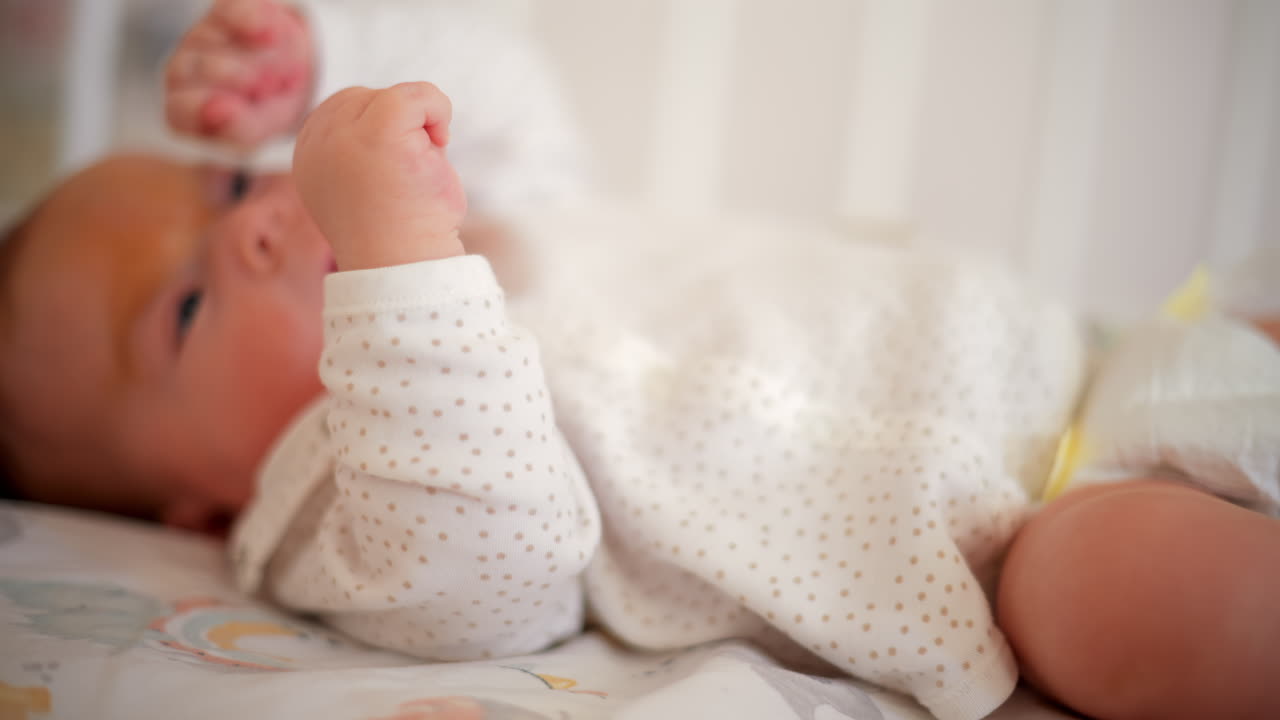 A newborn baby in soft pajamas moves gently in a crib, stretching tiny hands in natural morning light