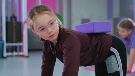 side view of a young girl with a purple block on her back during a gym class, another child partially visible and blurred in the background