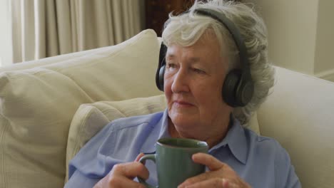 Mujer-Mayor-Caucásica-Usando-Audífonos-Escuchando-Música-Mientras-Sostiene-Una-Taza-De-Café-En-Casa