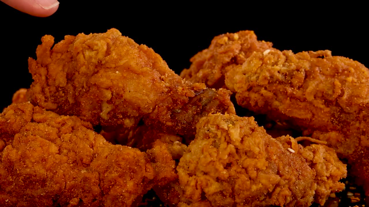 A hand reaches in to pick up a spicy, crumbed fried chicken piece from a pile, with dramatic lighting and slow-motion movement against a black background