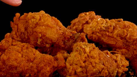 a hand reaches in to pick up a spicy, crumbed fried chicken piece from a pile, with dramatic lighting and slow-motion movement against a black background