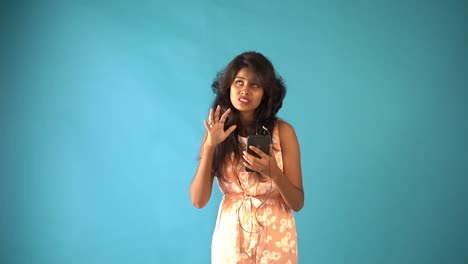 A-angry-young-Indian-girl-in-orange-frock-wearing-earphones-and-talking-in-video-chat-with-phone-in-an-isolated-blue-background