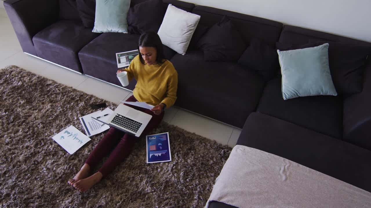 Mixed race woman sitting on floor using laptop going through paperwork drinking cup of coffee