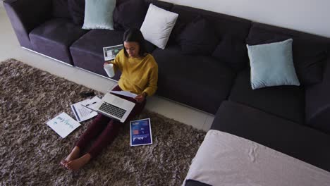 mixed race woman sitting on floor using laptop going through paperwork drinking cup of coffee