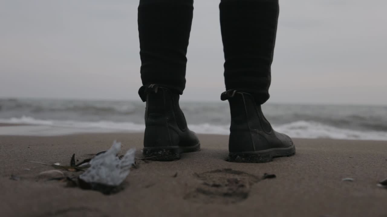 Un uomo in piedi sulla riva con scarpe nere e onde leggere che gli toccano le suole - Primo piano.