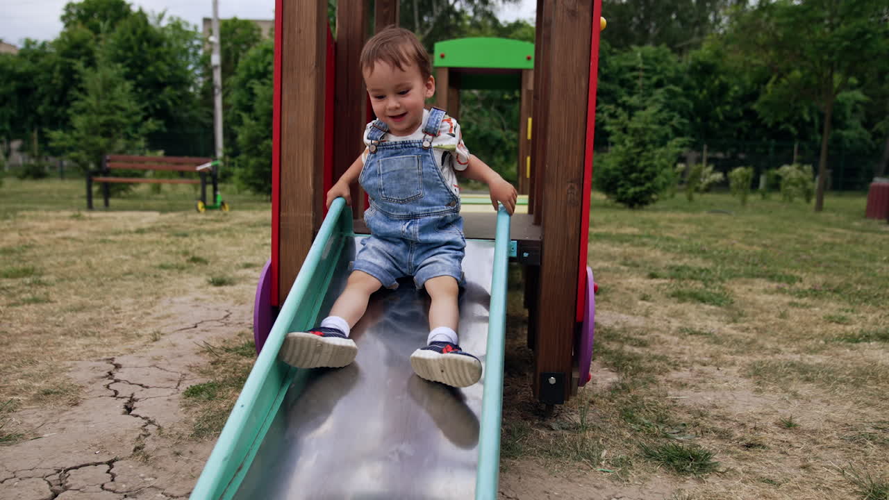 Beautiful baby boy in jeans romper sits on top of the slide. Kid hesitantly decides to go down smiling sweetly.