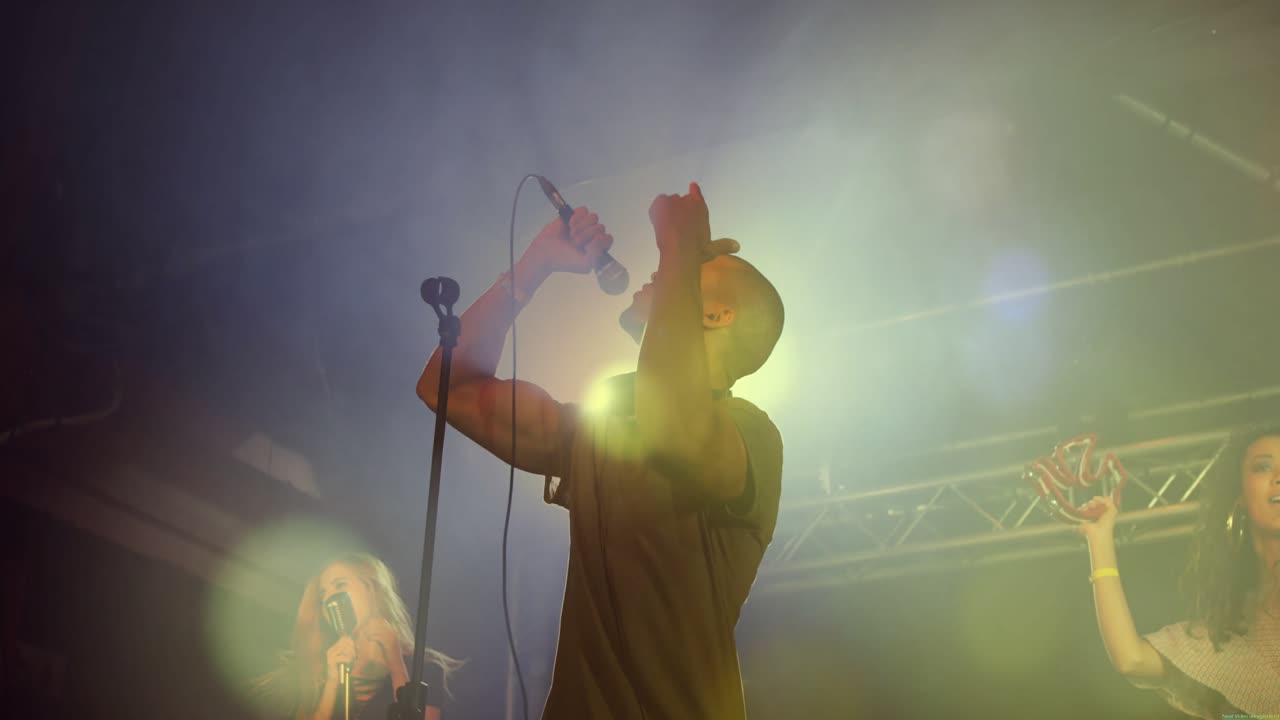 Cantante solista che avanza, afferrando il microfono e cantando, mentre i coristi ondeggiano sotto la foschia del concerto