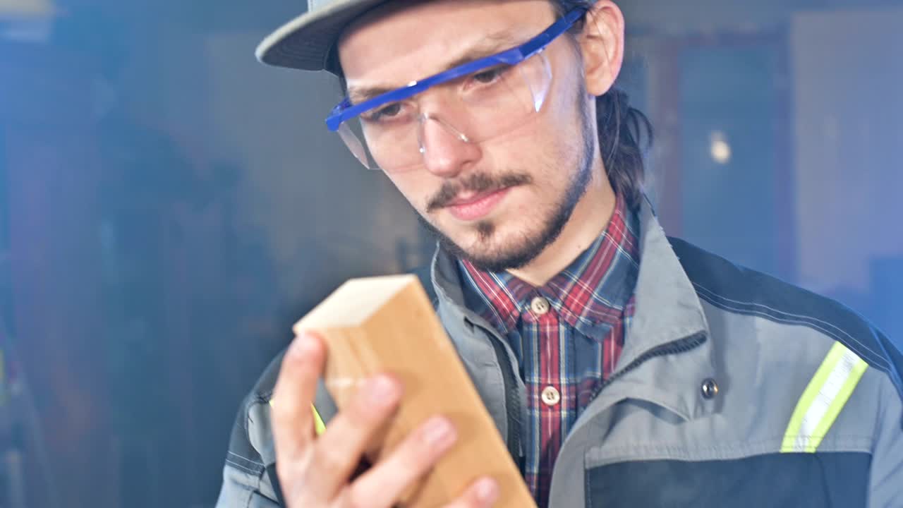 joven carpintero barbudo concentrado con gafas de protección y una gorra comprueba la pieza de trabajo