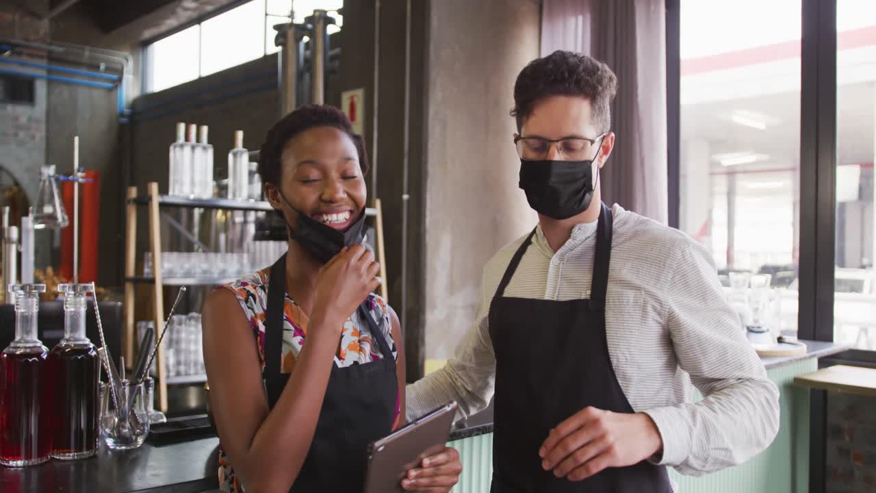 retrato de una pareja diversa con máscaras faciales trabajando en un bar, usando una tableta y sonriendo a la cámara