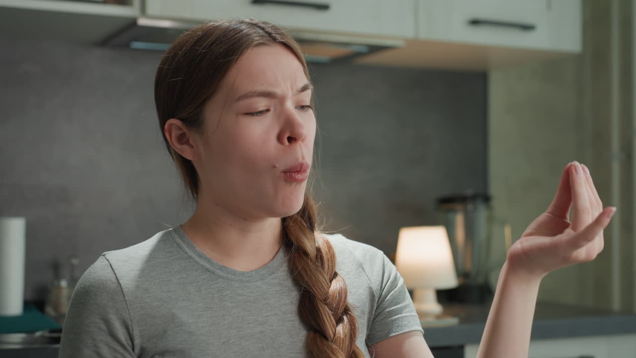 Mujer blanca embarazada probando especias y saboreando el aroma en una cocina moderna, pelo trenzado, camiseta informal, inhalación expresiva, momento de antojo juguetón, bienestar prenatal y ambiente de cocina casera.
