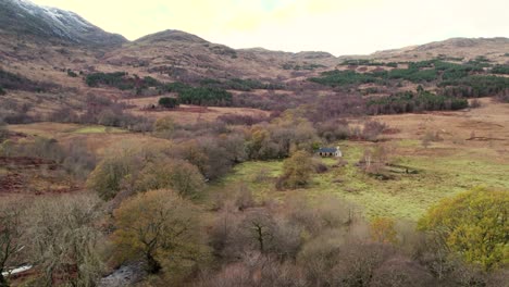 aerial drone footage slowly flying over trees in glen etive in the scottish highlands towards cadderlie bothy in winter set in a snow-capped mountain, native broadleaf woodland and a river landscape
