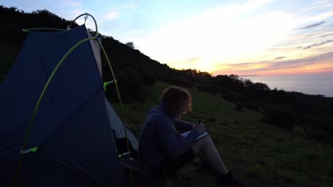 Un-Hombre-Escribiendo-Un-Cuaderno-En-Un-Camping-En-El-Océano-Al-Atardecer