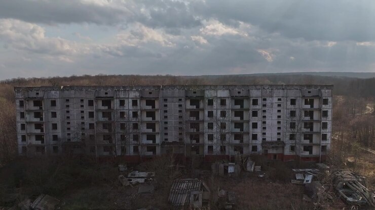 Vista de dron acercándose a un gran bloque de apartamentos abandonado en Moldavia, mostrando ventanas rotas, hormigón deteriorado y un paisaje postsoviético cubierto de vegetación bajo nubes dramáticas