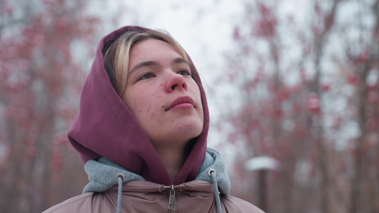 крупный план молодой женщины в зимней куртке с капюшоном, идущей вдумчиво и оглядывающейся вдумчиво, покрытый снегом парк с красными ягодными деревьями и мягким облачным небом