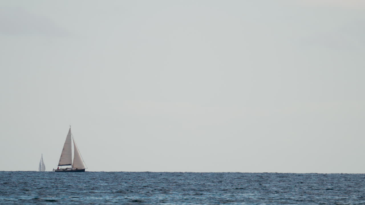 A white sailboat navigates through blue waves on a clear sunny day, with the horizon stretching endlessly behind it