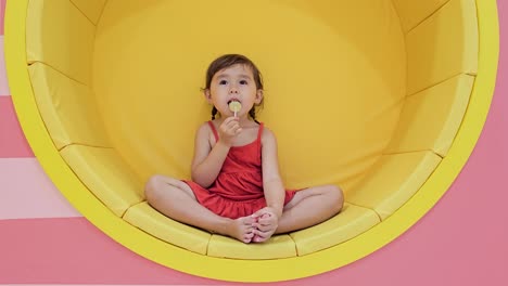 Cute-3-year-old-girl-lick-sweet-lollipop-candy-sitting-in-yellow-vivid-furniture-at-kid's-playroom