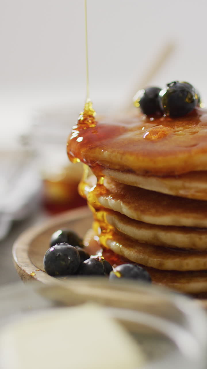 video vertical de jarabe de arce que se vierte en una pila de panqueques de estilo americano coronados con arándanos