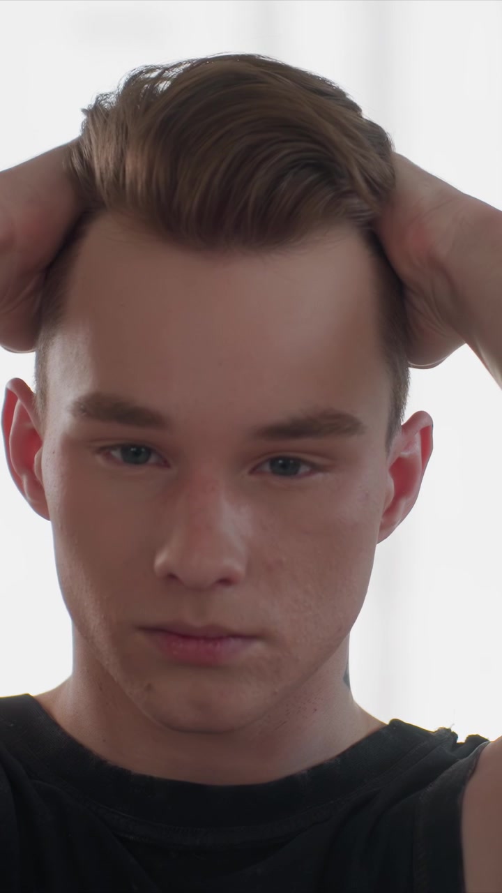 Primo piano di un giovane uomo che si sistema i capelli castani con movimenti ripetuti delle mani. L'uomo indossa una camicia nera e lo sfondo è di colore chiaro uniforme. Tatuaggio visibile sulla clavicola sinistra.