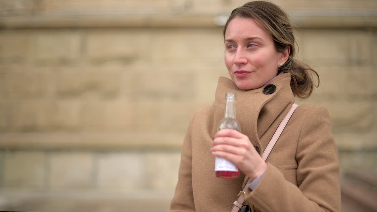 Woman in brown coat drinking a pink beverage outside