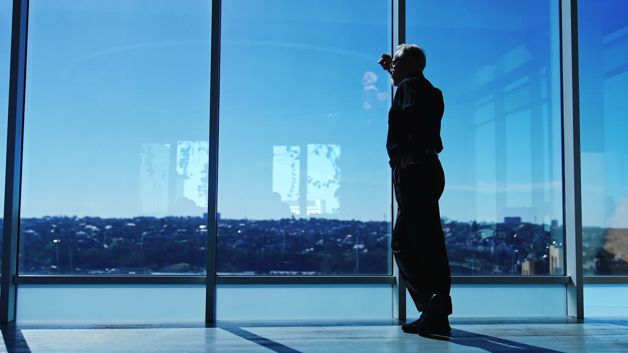 Pensive senior man stands leaning on the window frame. Man looks at the cityscape. Low angle view.