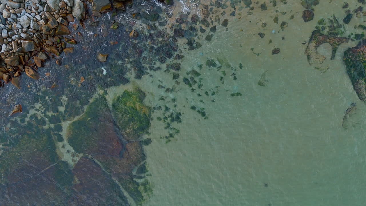Top down aerial view of rocky sea bottom and clear water. Overhead drone shot of shallow sea water with visible green mossy rocks and sandy bottom