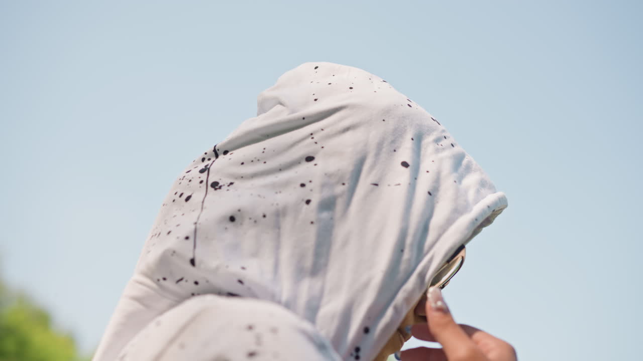 검은색 후드티를 입은 십 대가 야외에서 흰색 후드티를 조절하고 있어요. 햇빛을 가리기 위해 안경 위로 후드를 잡아당기는 손, 면직물에 묻은 페인트 얼룩 디테일, 집중된 자세, 솔직한 거리 분위기가 공원 샷에 담겨 있어요.